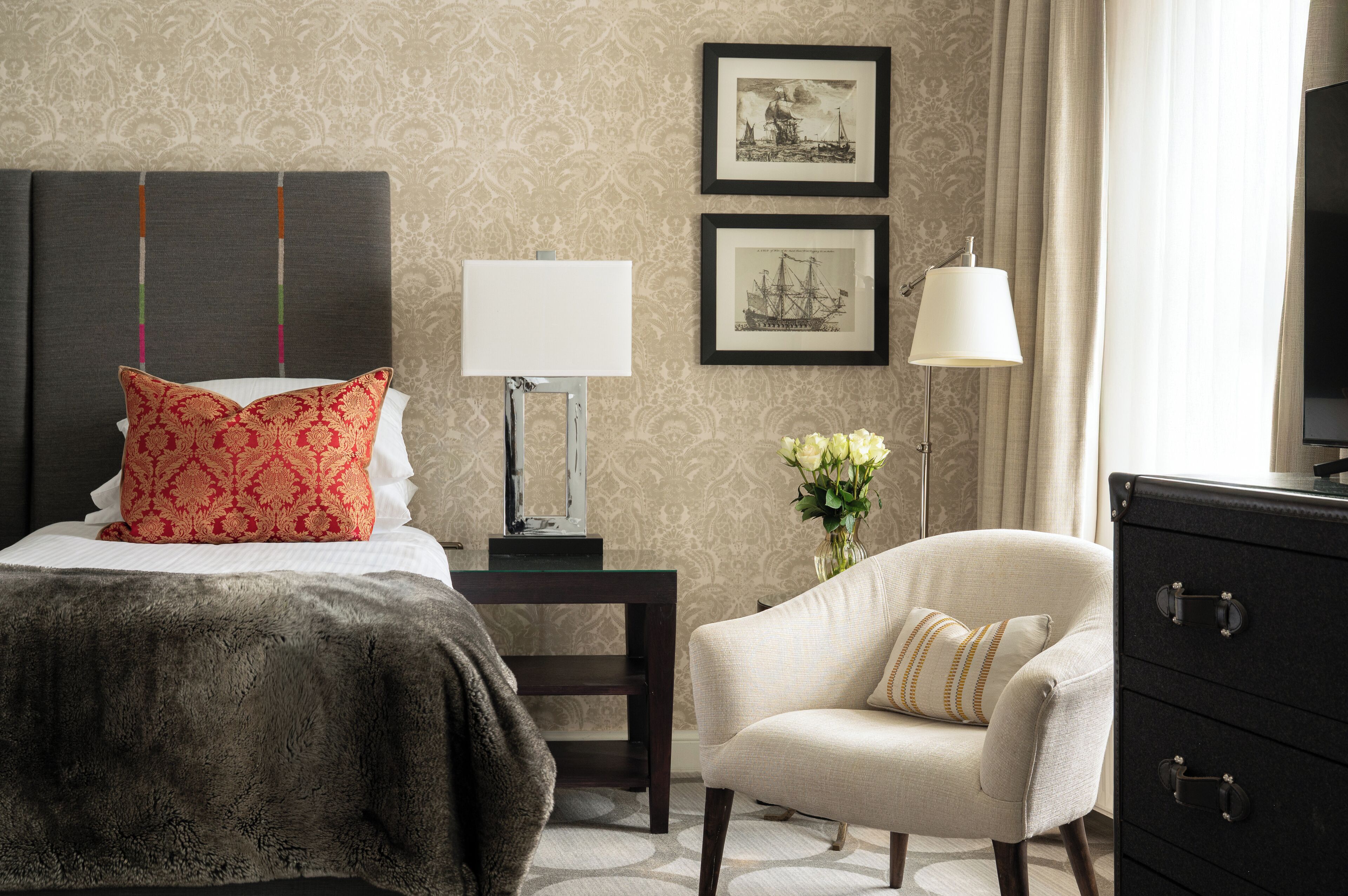A hotel room with a bed, a chair, a nightstand, a lamp, a dresser, and two framed pictures on the wall.