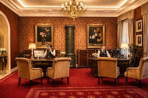 Una sala formal con alfombra roja, dos escritorios de madera, sillas, un jarrón grande y cuadros en las paredes.
