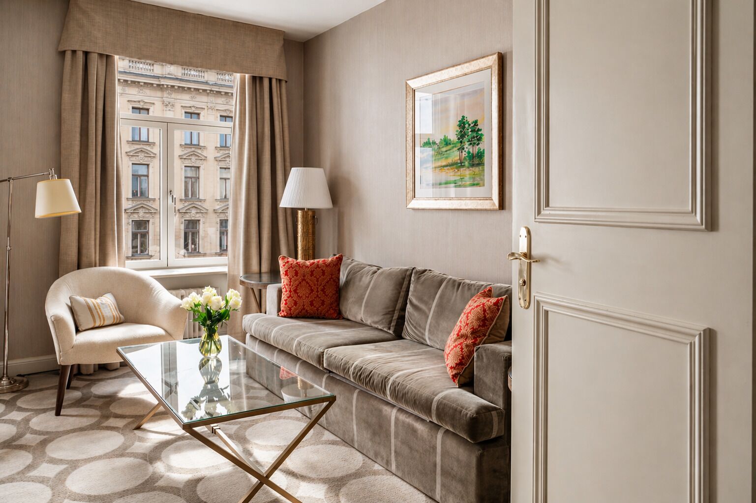 A hotel room with a sofa, armchair, glass coffee table, and a view of a building through large windows.