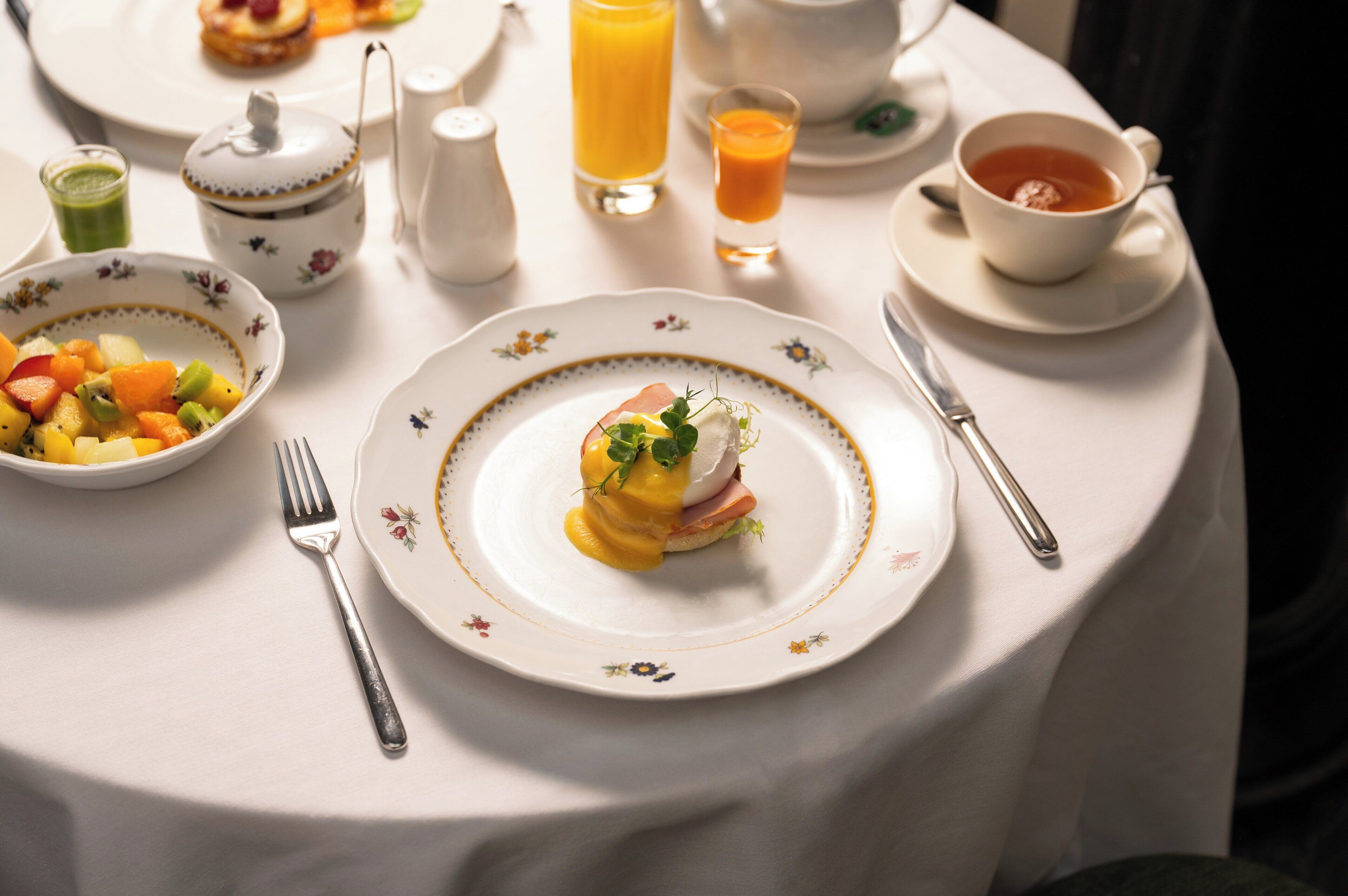 Un desayuno con un plato de huevos benedictinos, un bol de fruta y una taza de té sobre una mesa blanca.