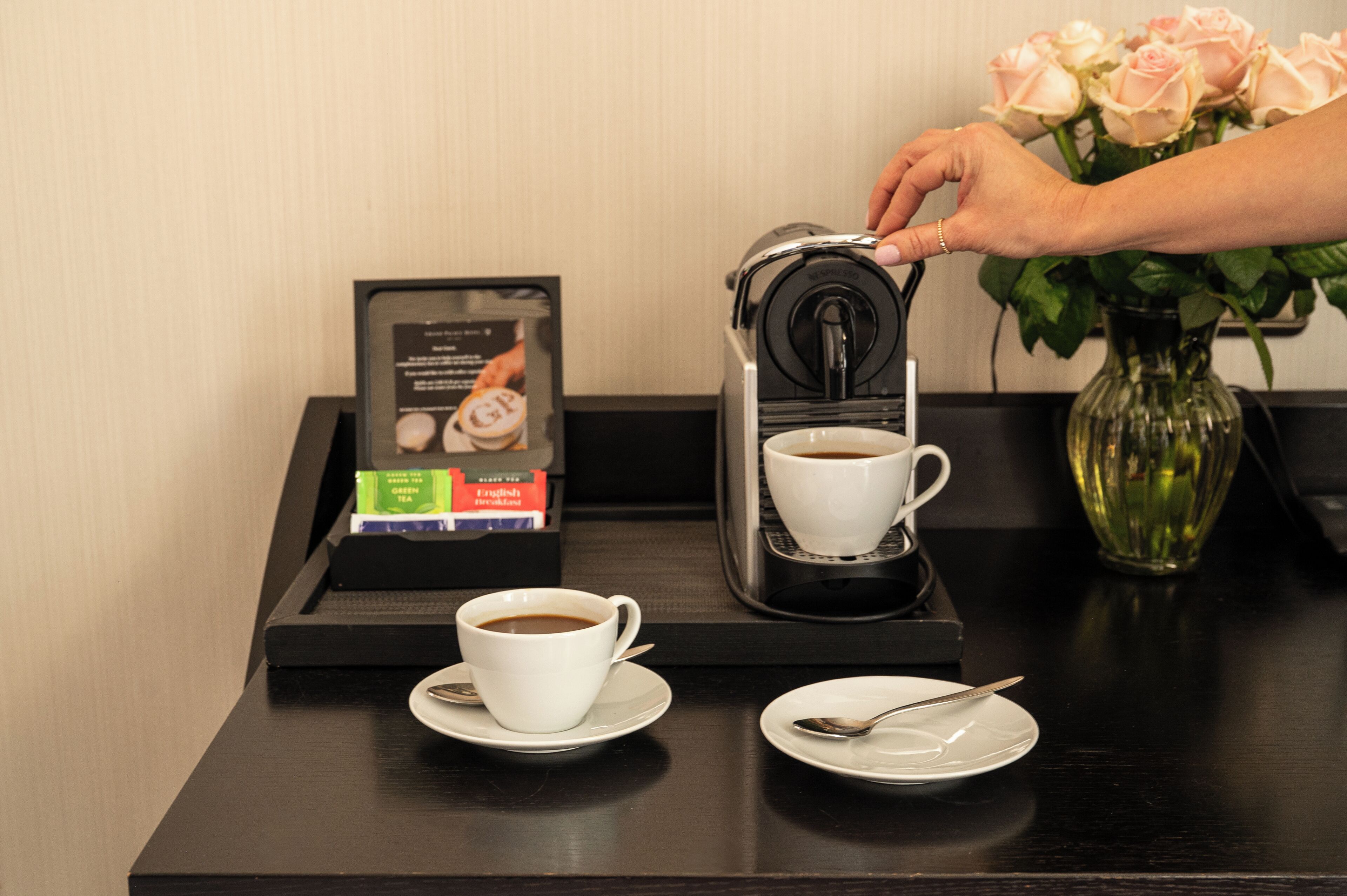A coffee machine dispensing coffee into a cup, a tea box, a vase with roses, and a menu on a tray.