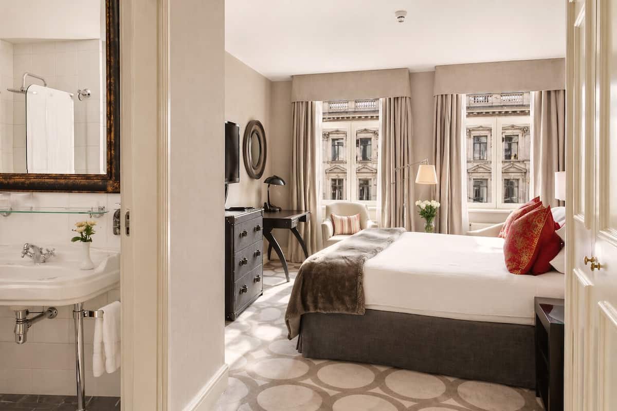 Habitación de hotel con cama, cómoda, tocador con espejo y ventana con cortinas, con vista a un edificio.