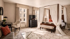 A hotel room with a four-poster bed, a TV, a desk with a lamp, a chair, and a view of the city. - Grand Palace Hotel – Small Luxury Hotels of the World (Riga)