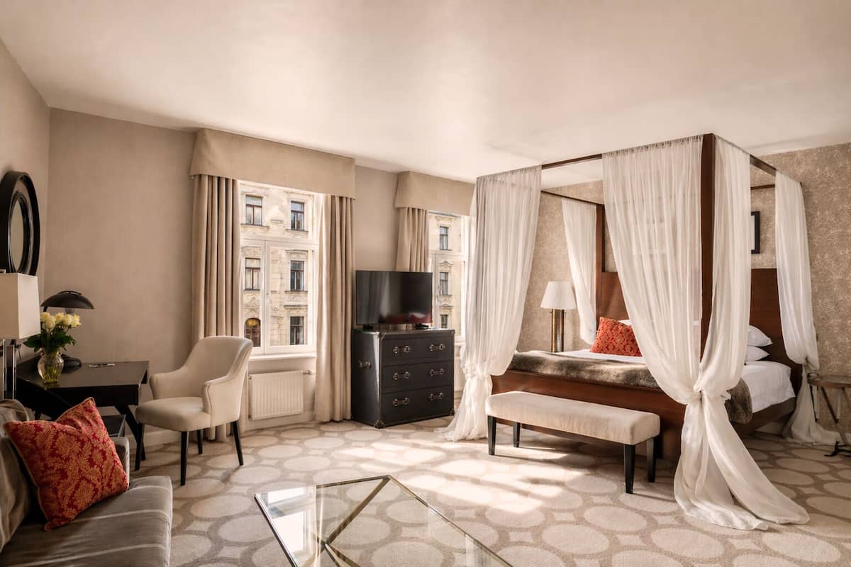 Habitación de hotel con cama con dosel, televisión, escritorio con lámpara, una silla y vistas a la ciudad.