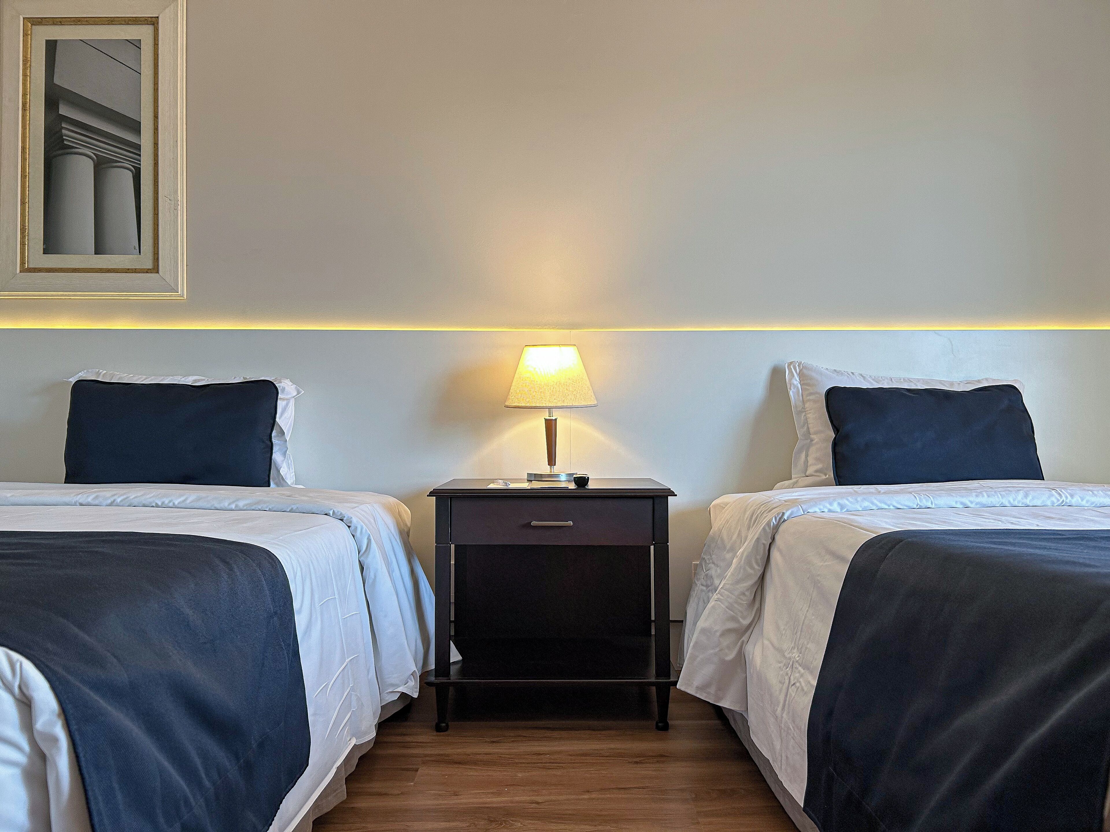 Quarto de hotel com duas camas, uma mesa de cabeceira com abajur e um quadro na parede.