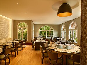 A spacious dining area with multiple tables and chairs, large arched windows, and a warm ceiling light.