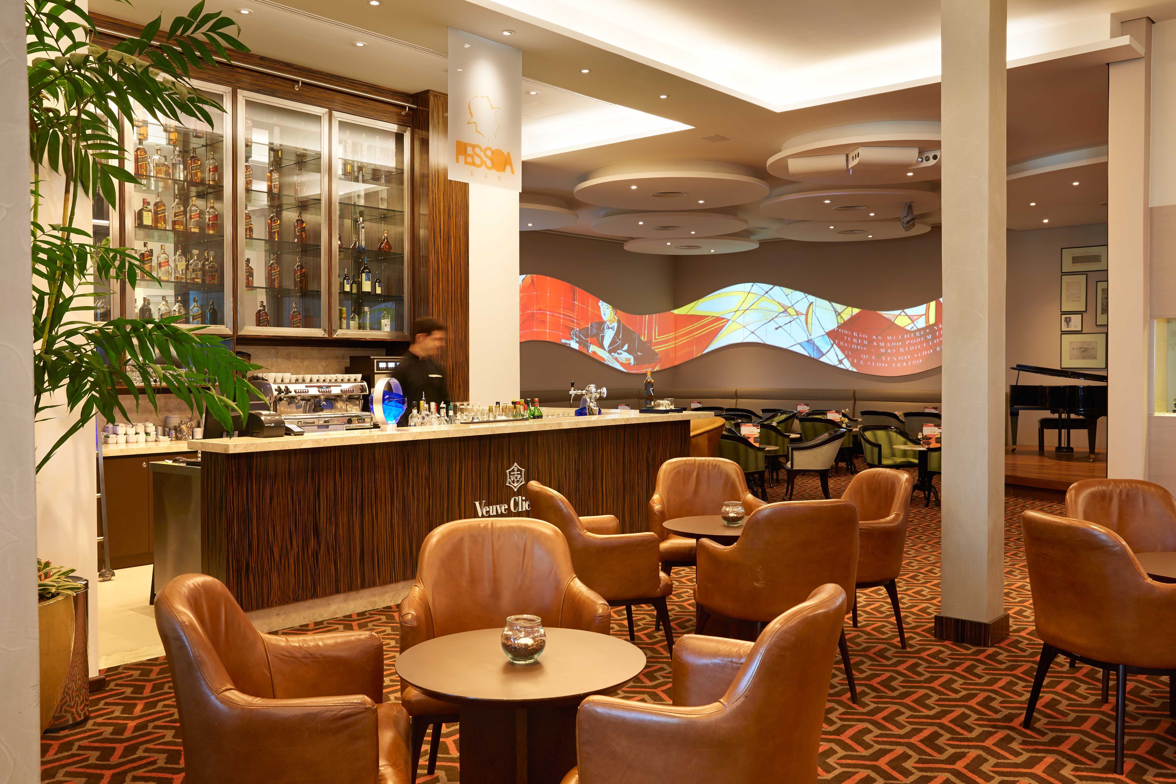 Zona de bar con sillones de cuero y mesas, un minibar y una pantalla grande que muestra a una persona.