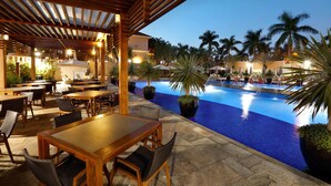 An outdoor pool area with wooden tables and chairs, surrounded by palm trees and potted plants. - Royal Palm Plaza Resort Campinas (Campinas)
