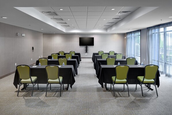 A conference room with multiple tables and chairs, a flat-screen TV mounted on the wall, and large windows with curtains.
