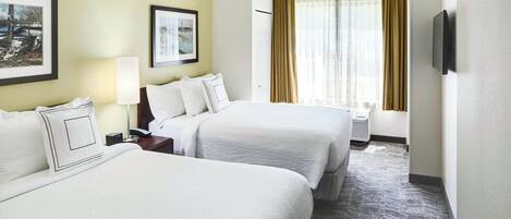 A hotel room with two beds, a bedside table with a lamp, a TV mounted on the wall, and a window with curtains.