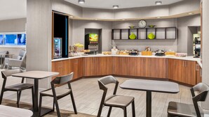 Una cocina moderna con armarios de madera, un refrigerador empotrado y una encimera con diversos alimentos. Hay varias sillas y mesas dispuestas alrededor de la cocina.