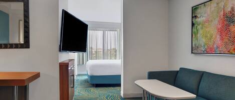 Ein Hotelzimmer mit einem Bett, einem an der Wand befestigten Fernseher, einem kleinen Tisch und einem farbenfrohen, abstrakten Gemälde.