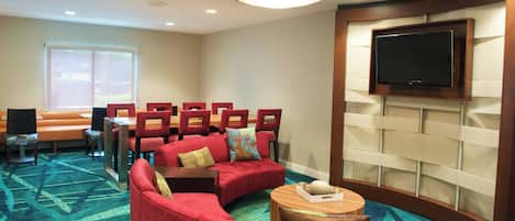 A hotel room with a red sofa, a small round table, a flat-screen TV mounted on the wall, and a large window with blinds.