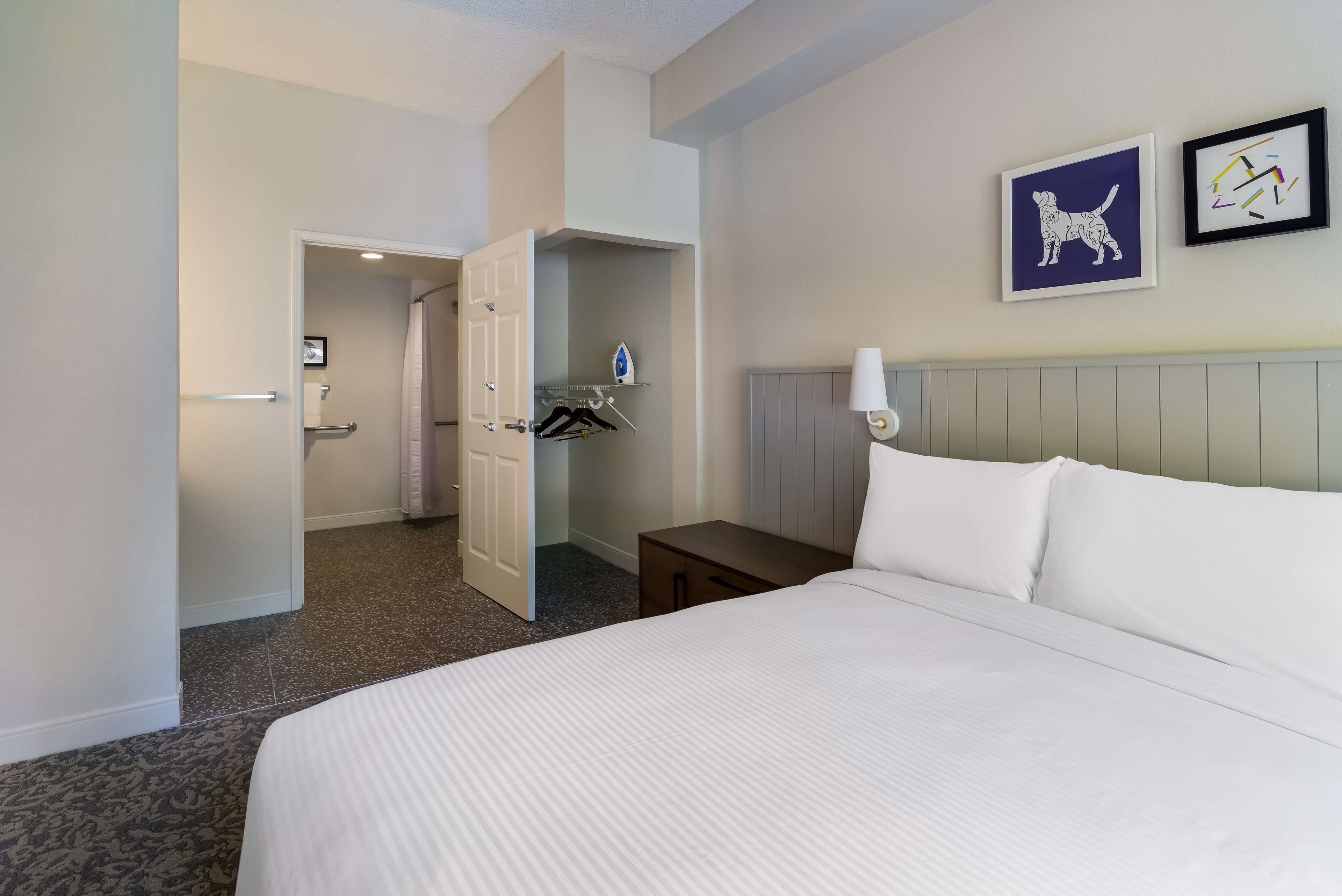 A hotel room with a bed, a nightstand, a wall-mounted lamp, a door leading to a bathroom, and two framed pictures on the wall.