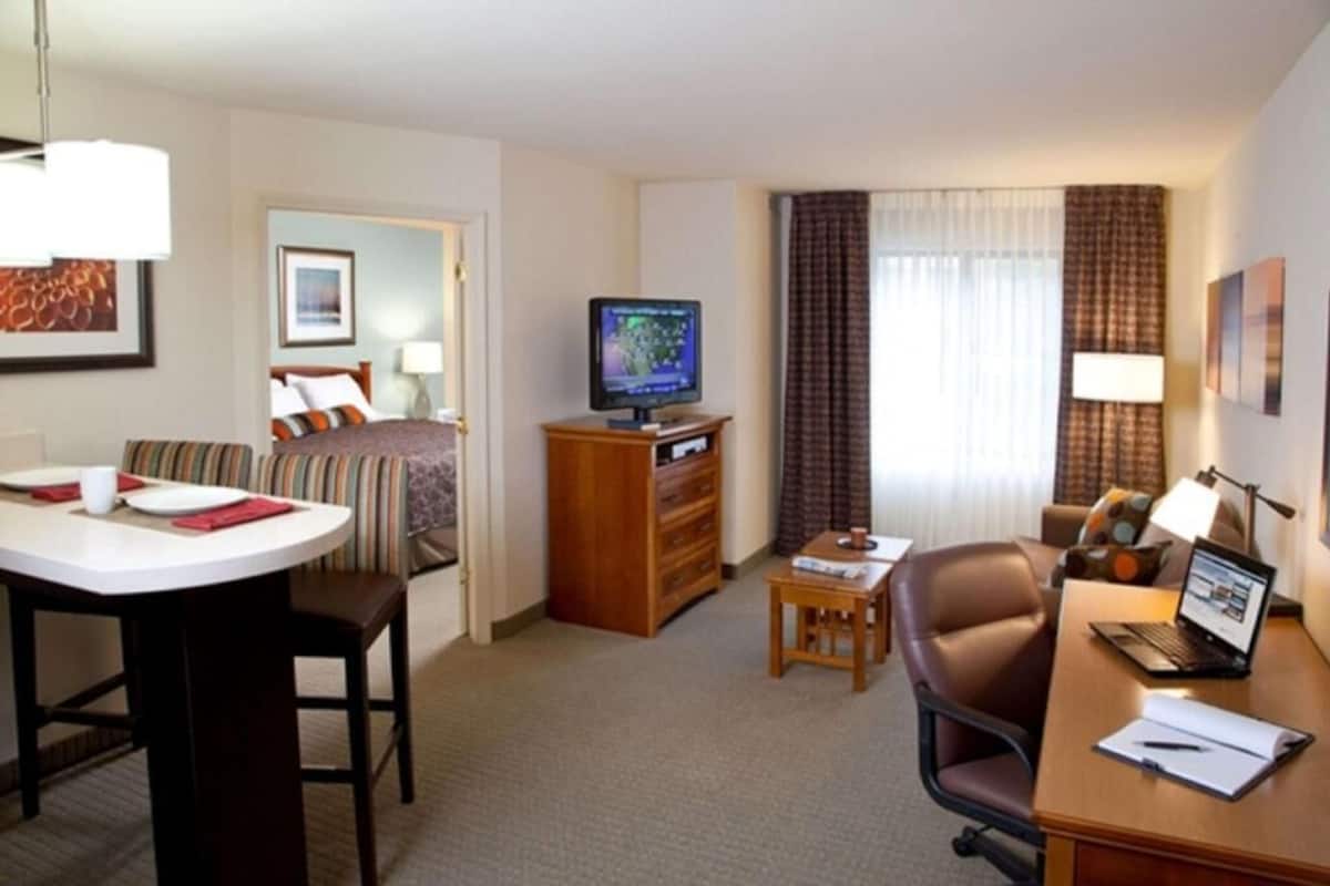 Una habitación de hotel con cama, televisión, escritorio con una computadora portátil, una silla y una mesita.