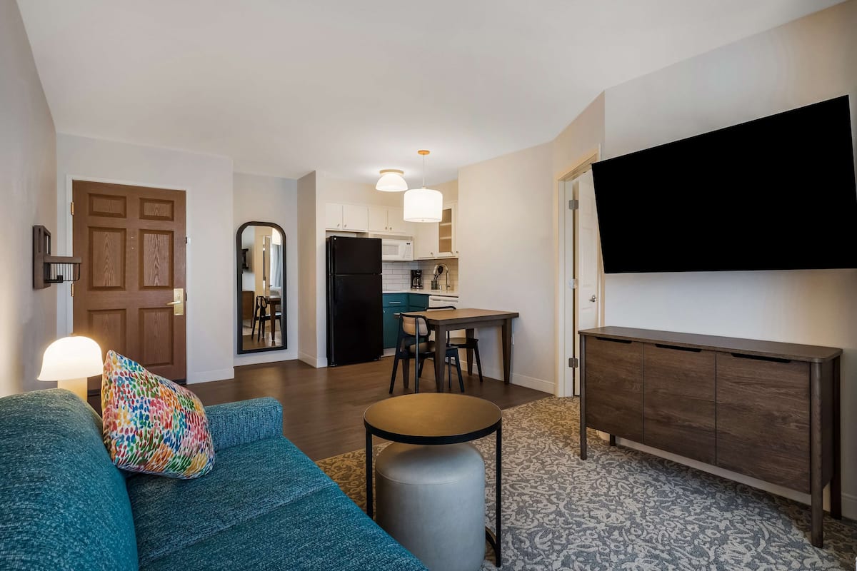 Una sala de estar moderna con un sofá color turquesa, una mesita redonda pequeña y un mueble bajo de madera oscura. Se aprecia, al fondo, una cocina con armarios blancos y un refrigerador negro.