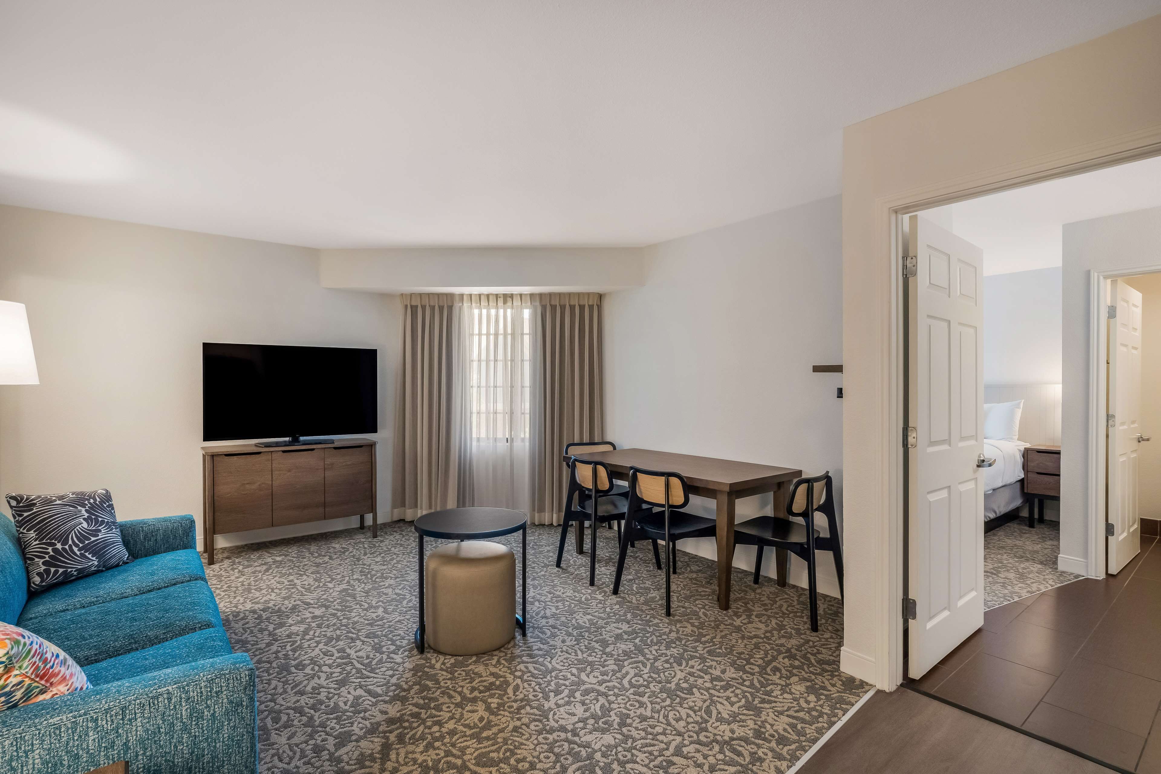 A hotel room with a sofa, a TV, a dining table with chairs, a small ottoman, and a patterned carpet.