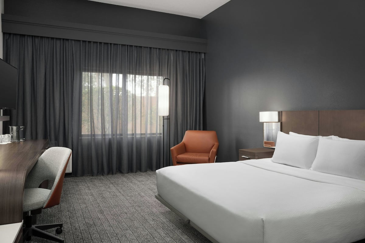 Een hotelkamer met een groot bed, een bureau met stoel, een nachtkastje met lamp en een raam met gordijnen.