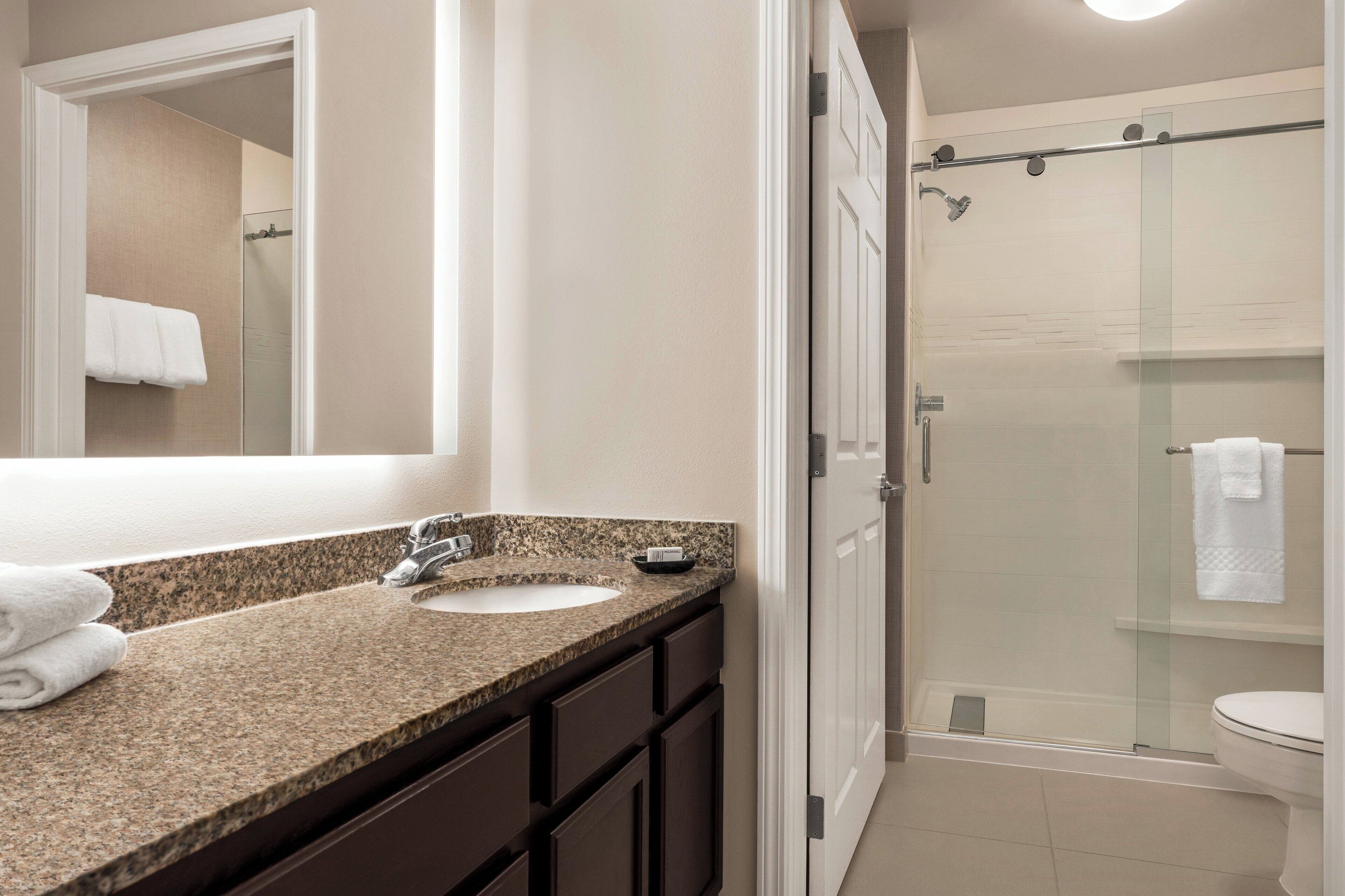 A bathroom with a granite countertop sink, dark cabinets, a large mirror, and a glass-enclosed shower.
