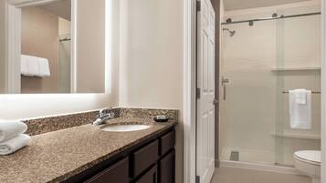 A bathroom with a granite countertop sink, dark cabinets, a large mirror, and a glass-enclosed shower.