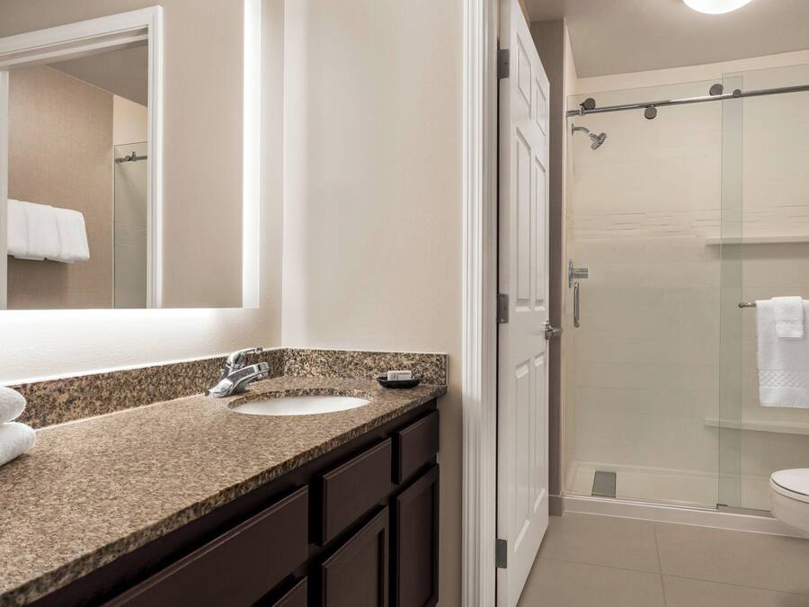 A bathroom with a granite countertop sink, dark cabinets, a large mirror, and a glass-enclosed shower.