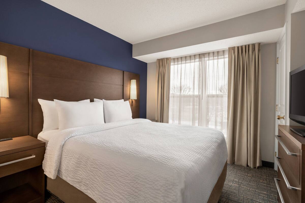 Habitación de hotel con una cama grande, un cabecero de madera, un televisor y una ventana con cortinas.