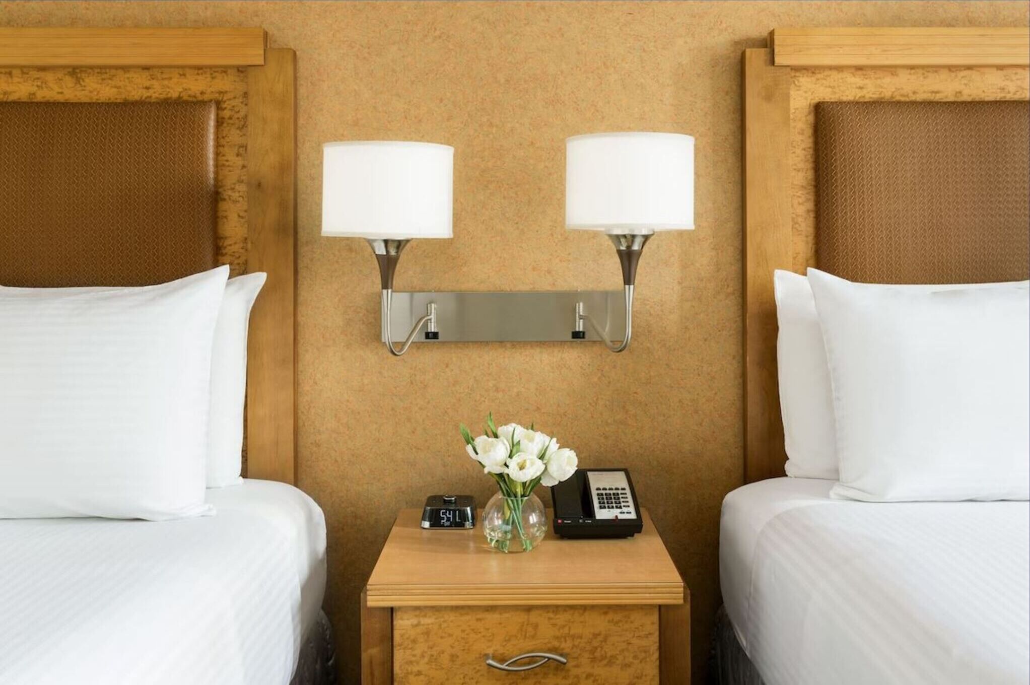 A hotel room with two beds, a nightstand with a clock and phone, and wall-mounted lamps.
