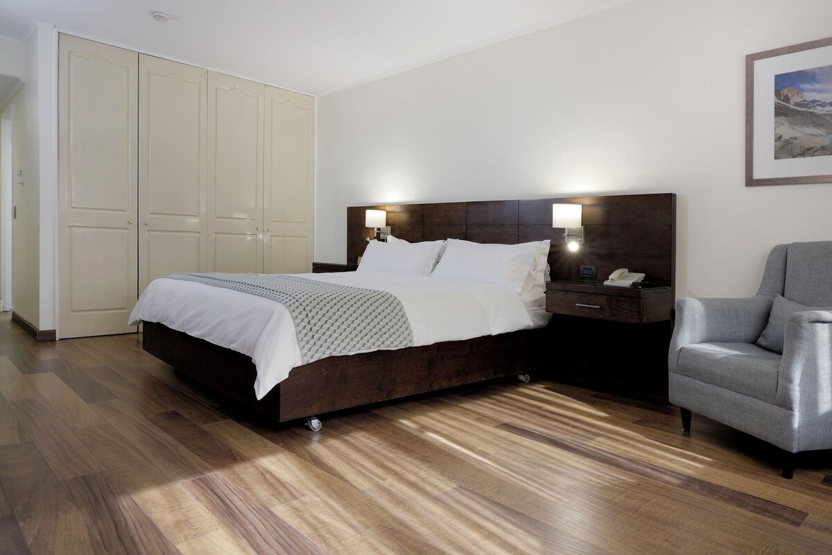 Habitación de hotel con cama, mesita de noche, silla, armario y un cuadro enmarcado en la pared.