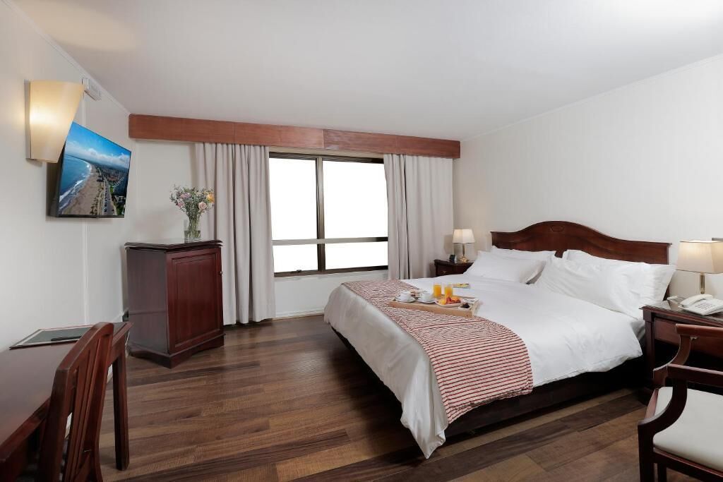 Habitación de hotel con cama, mesitas de noche, escritorio, silla, ventana con cortinas, televisor montado en la pared y un pequeño armario.