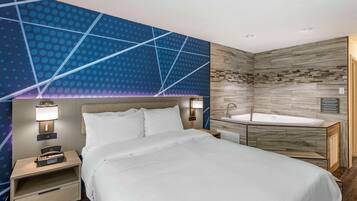 Quarto de hotel moderno com uma cama grande, uma mesa de cabeceira com luminária, uma banheira integrada e uma parede decorativa com padrão geométrico.