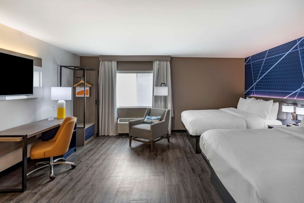 Une chambre d’hôtel équipée d’un bureau, de deux lits, d’une télévision, d’une chaise et d’une fenêtre avec des rideaux.