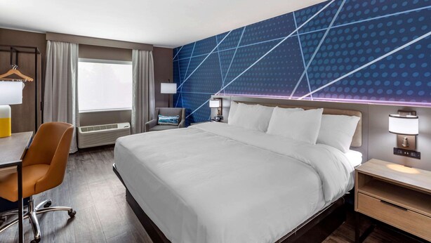 A hotel room with a large bed, a desk with a chair, a nightstand, and a wall-mounted lamp.