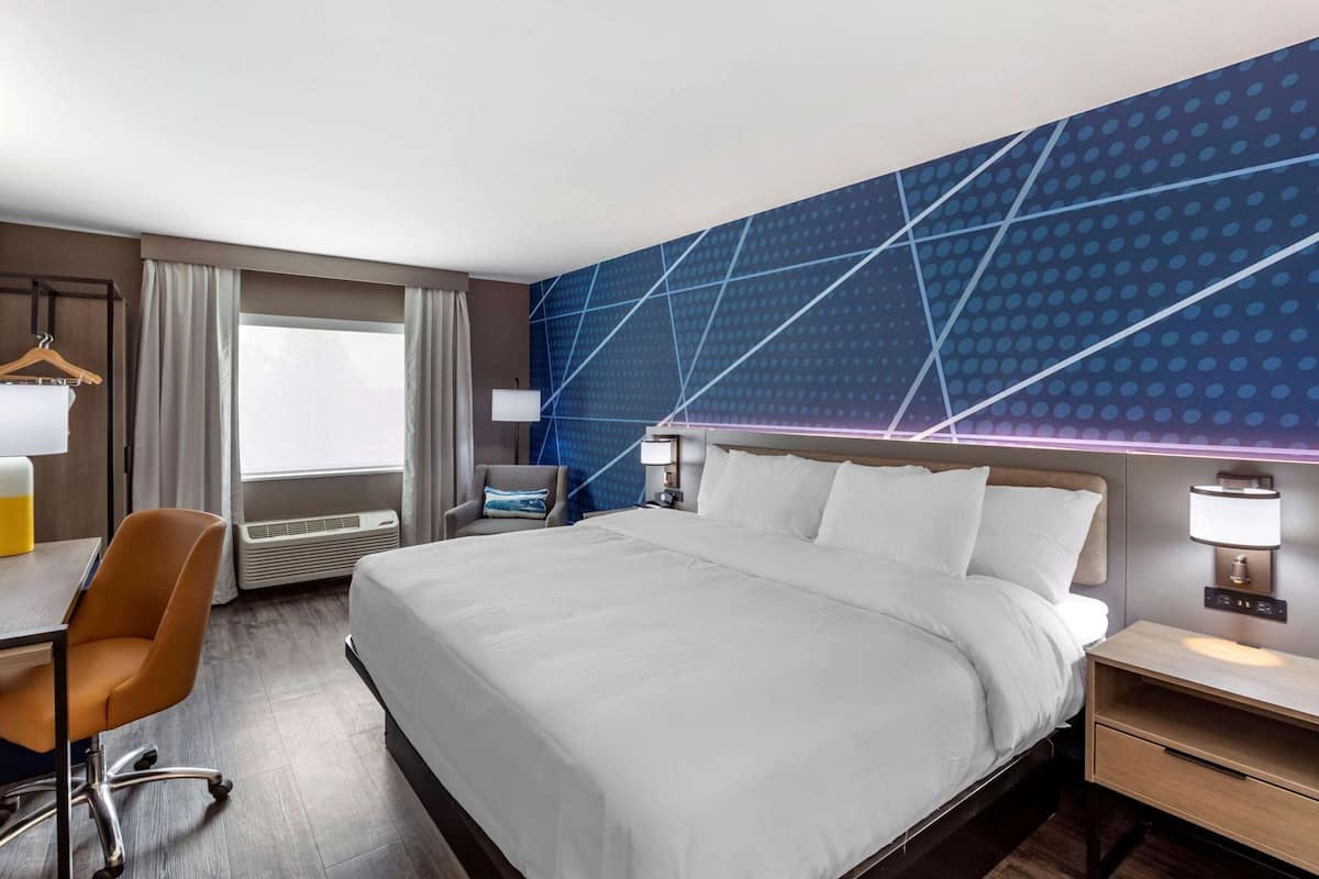 Une chambre d’hôtel avec un grand lit, un bureau, une chaise, une table de chevet et un mur orné d’un motif géométrique.