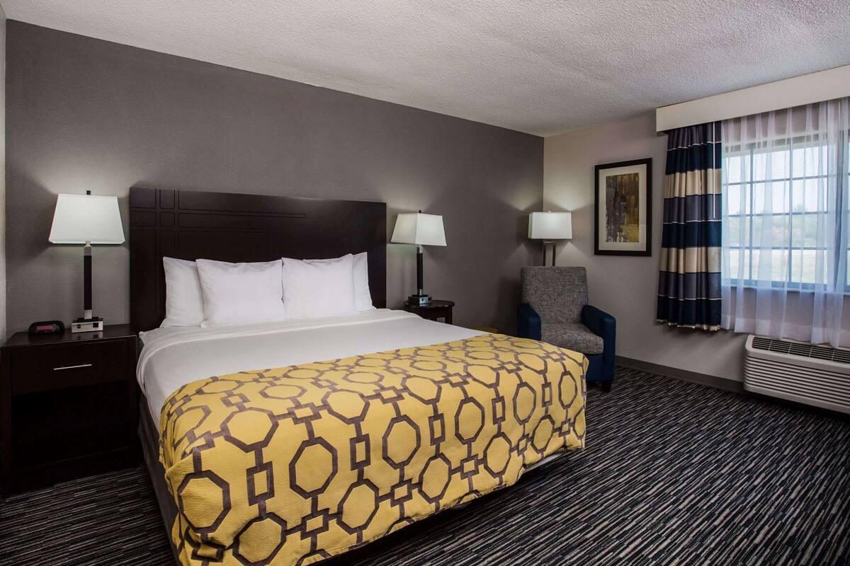 A hotel room with a large bed, two bedside tables with lamps, a chair, a window with curtains, and a wall-mounted air conditioner.