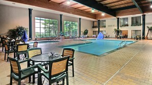Piscina interna com piso ladrilhado, vigas de madeira no teto e várias cadeiras e mesas ao redor da área da piscina.