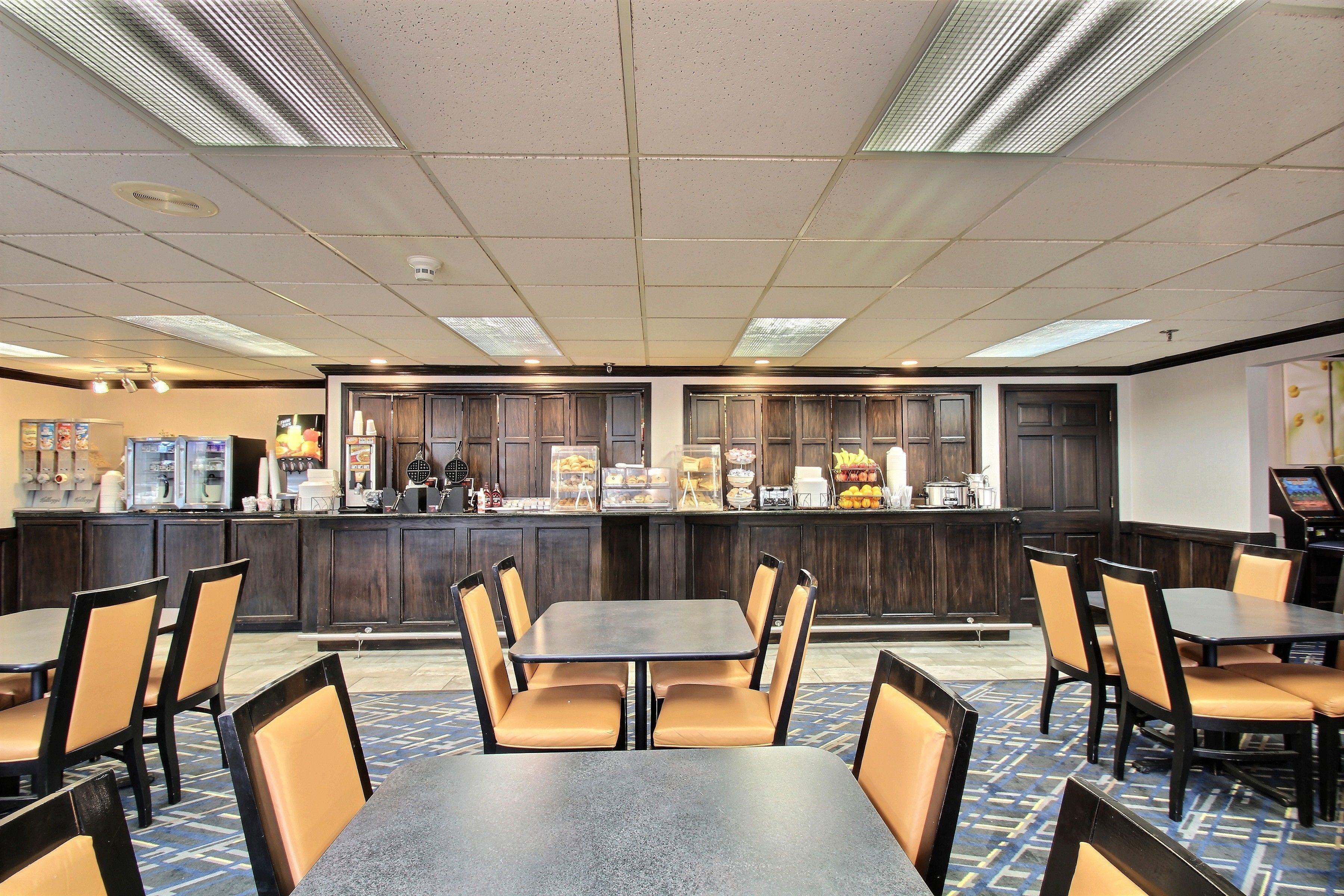 A hotel lobby with multiple tables and chairs, a counter with food and beverage options, and a patterned floor.