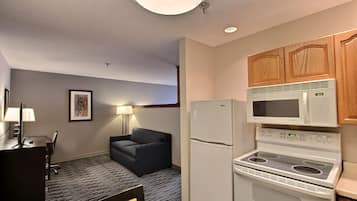 A compact hotel room with a kitchenette including a microwave, refrigerator, and stove. There is a desk with a chair and a sofa.
