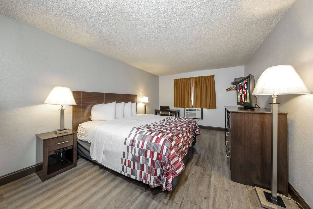 A hotel room with a large bed, two bedside tables with lamps, a wooden headboard, a desk with a chair, a wardrobe, and a television on a stand.