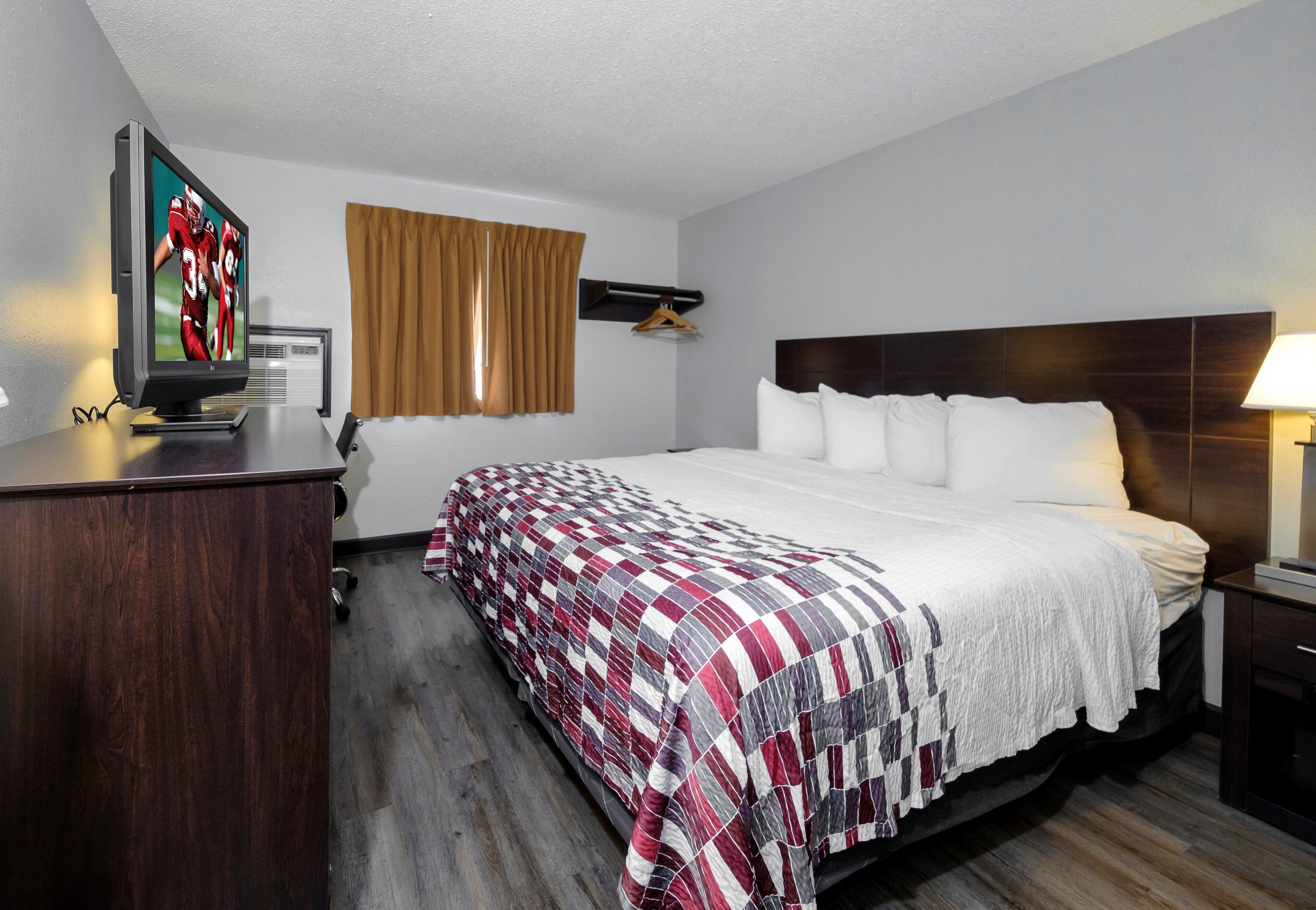 Una habitación de hotel con cama, escritorio con televisor, ventana con cortinas y mesita de noche con lámpara.