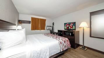 A hotel room with a bed, a flat-screen TV, a desk with a computer, a lamp, and a window with curtains.