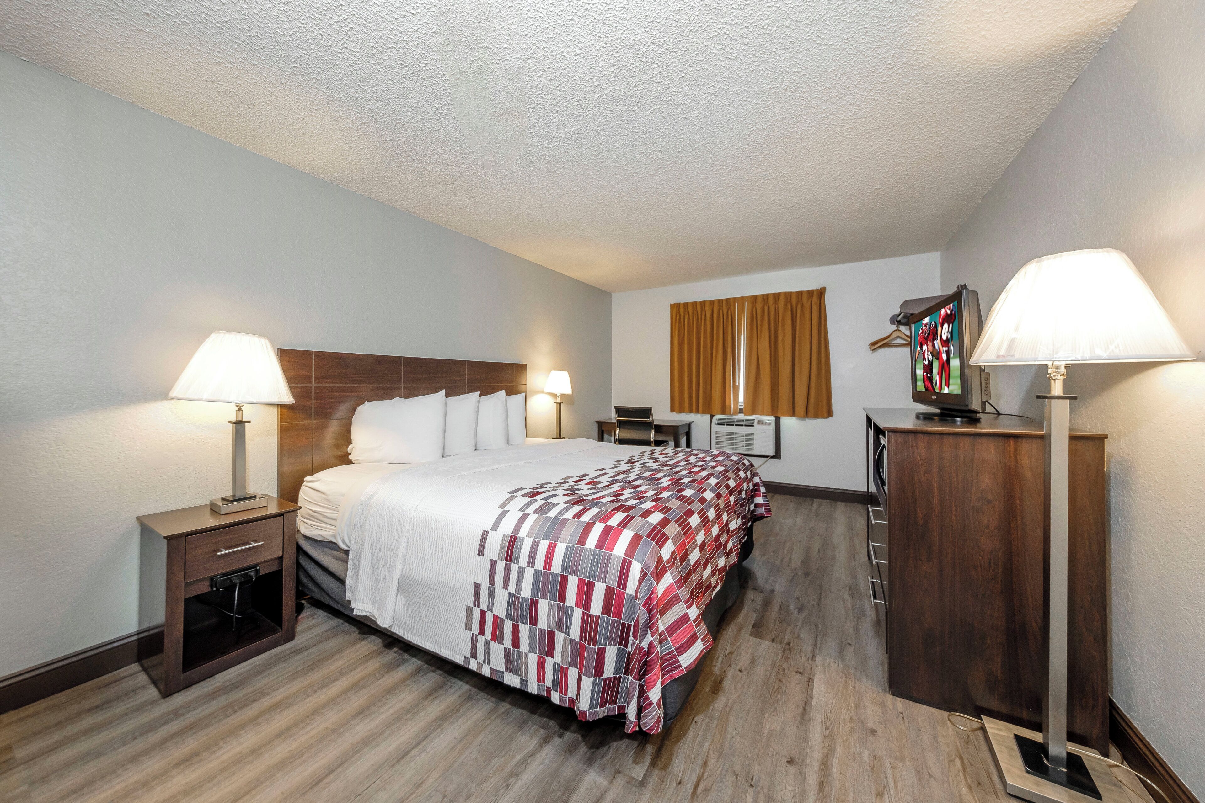 A hotel room with a bed, two bedside lamps, a desk, a chair, a TV, and a wardrobe.