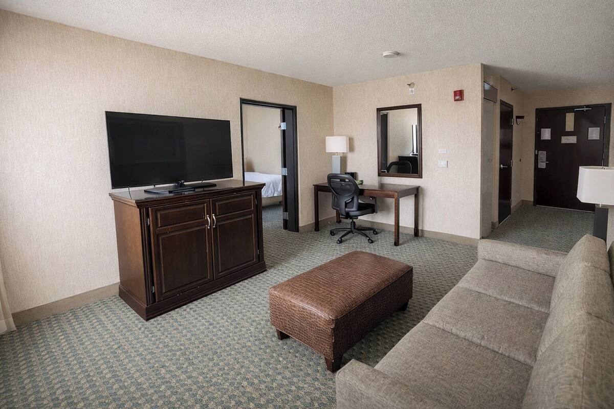 A hotel room with a television, a desk with a chair, a sofa, and a small ottoman.
