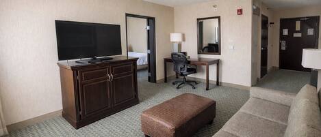 A hotel room with a television, a desk with a chair, a sofa, and a small ottoman.