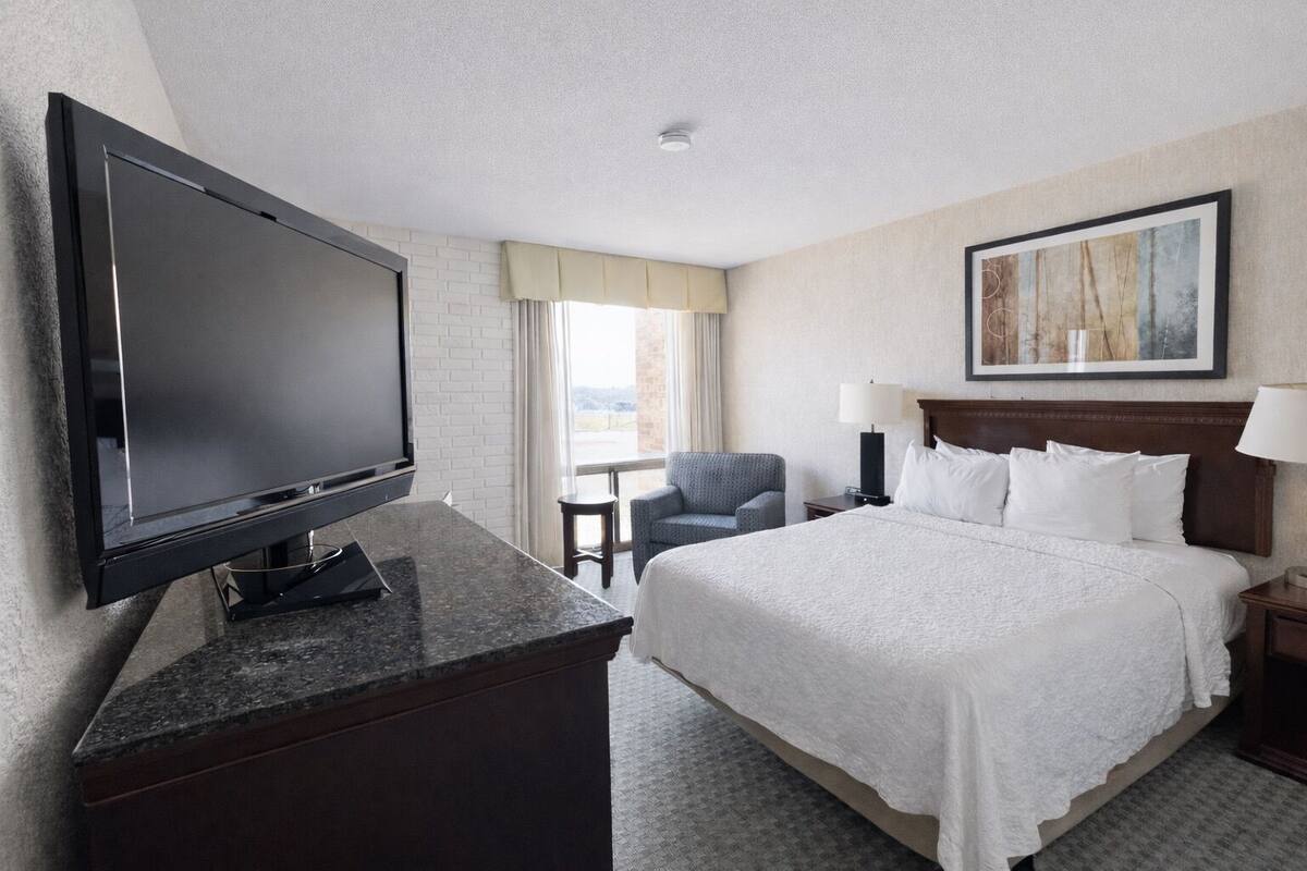 A hotel room with a bed, a TV, a chair, a lamp, and a painting on the wall.