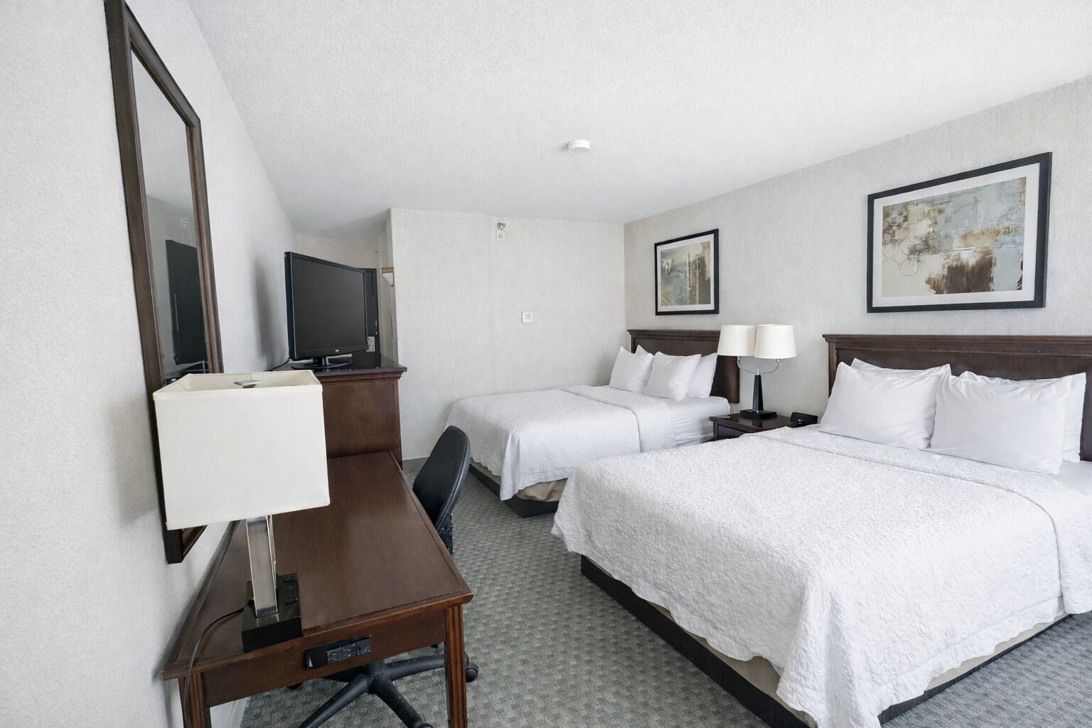 A hotel room with two beds, a desk with a chair, a TV, and two bedside tables with lamps.
