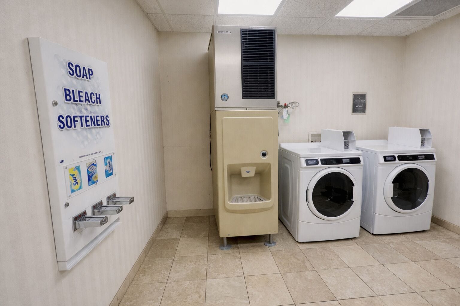 A laundry room with a wall-mounted soap, bleach, and softeners dispenser, a water heater, and two front-loading washing machines.