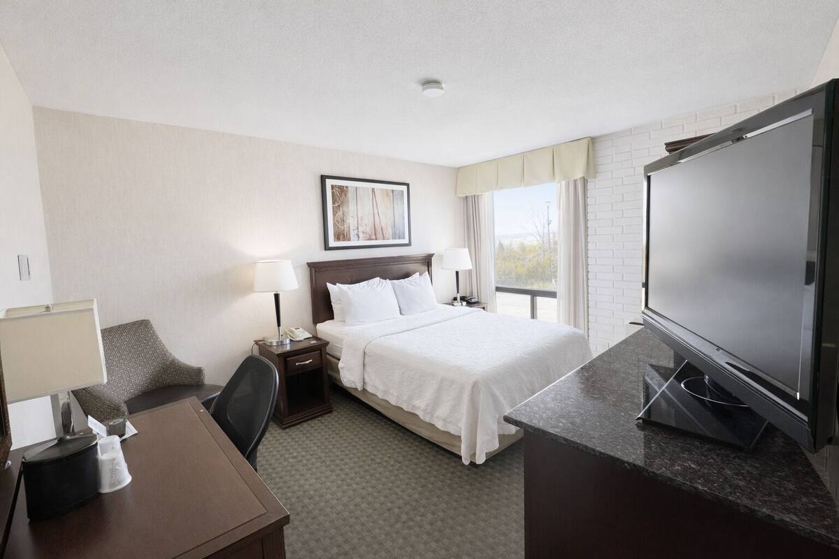 A hotel room with a bed, a desk, a chair, a TV, and a window with curtains.