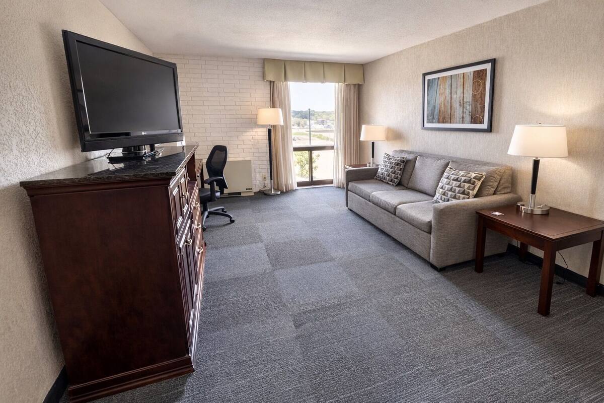 A hotel room with a flat-screen TV, a sofa, a desk, a dresser, and a window with curtains.