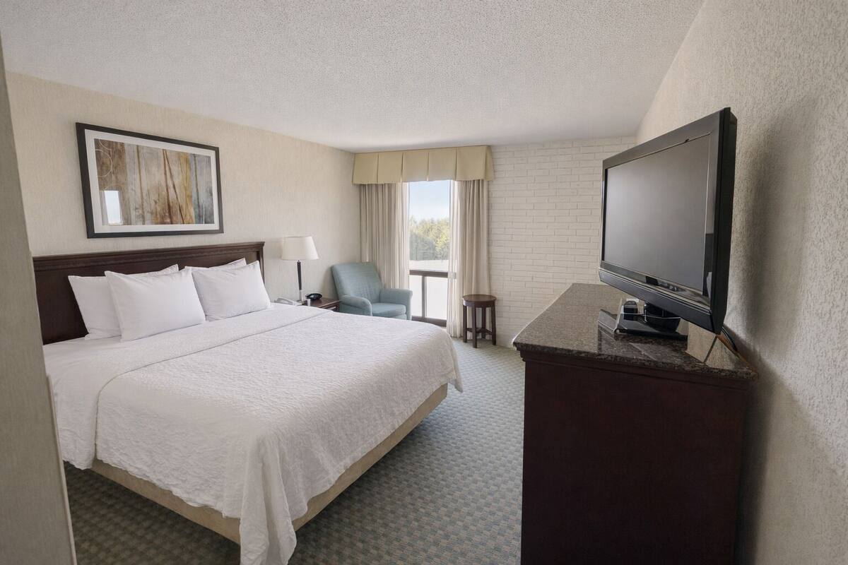 A hotel room with a large bed, a television on a desk, a chair, a small table, and a window with curtains.