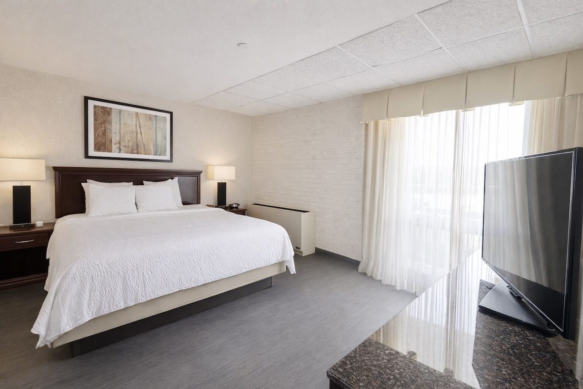 A hotel room with a large bed, a flat-screen TV, a painting on the wall, and a window with curtains.